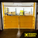 Interior view of a yellow self-closing mezzanine gate guarding an open roll-up doorway with stored pallets beyond.