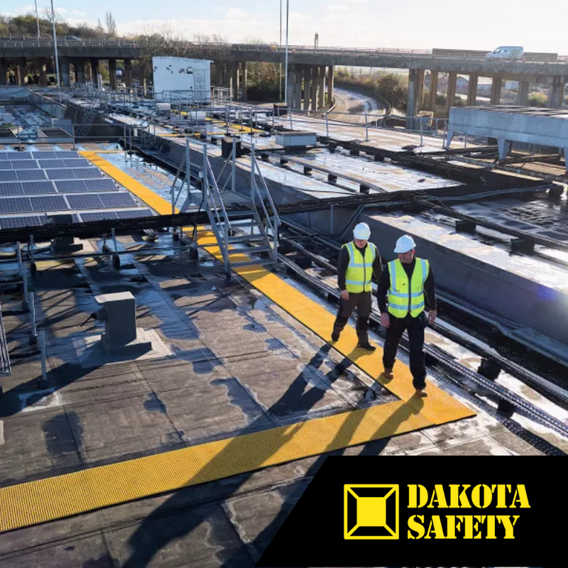Two workers in high-visibility vests and hard hats walking on yellow safety walkways installed between solar panels on a commercial rooftop, with Dakota Safety logo in bottom corner